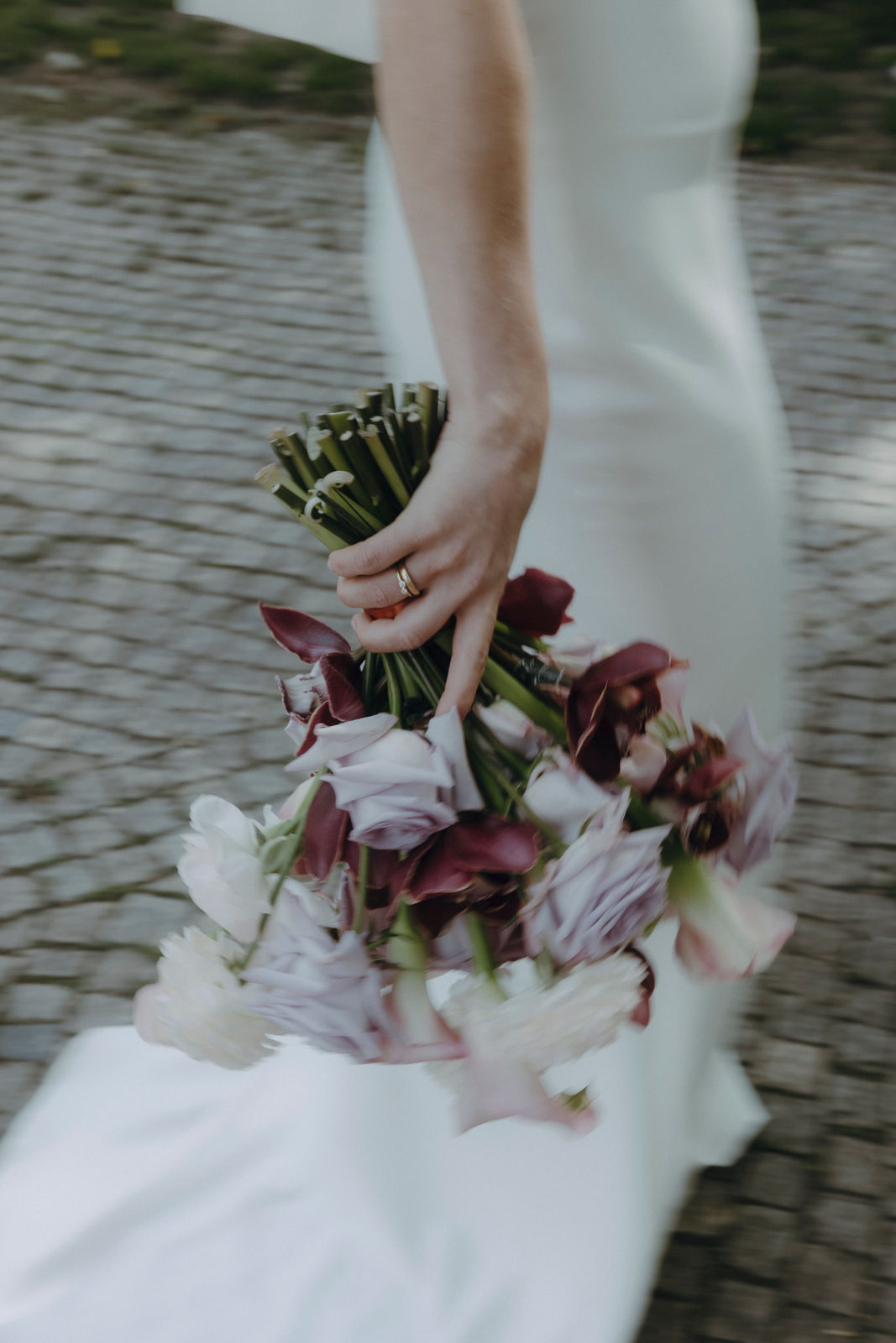 Bildausschnitt einer Braut. Man sieht die Hand mit Ehering und Verlobungsring, den Brautstrauß in Lilatönen und das Brautkleid